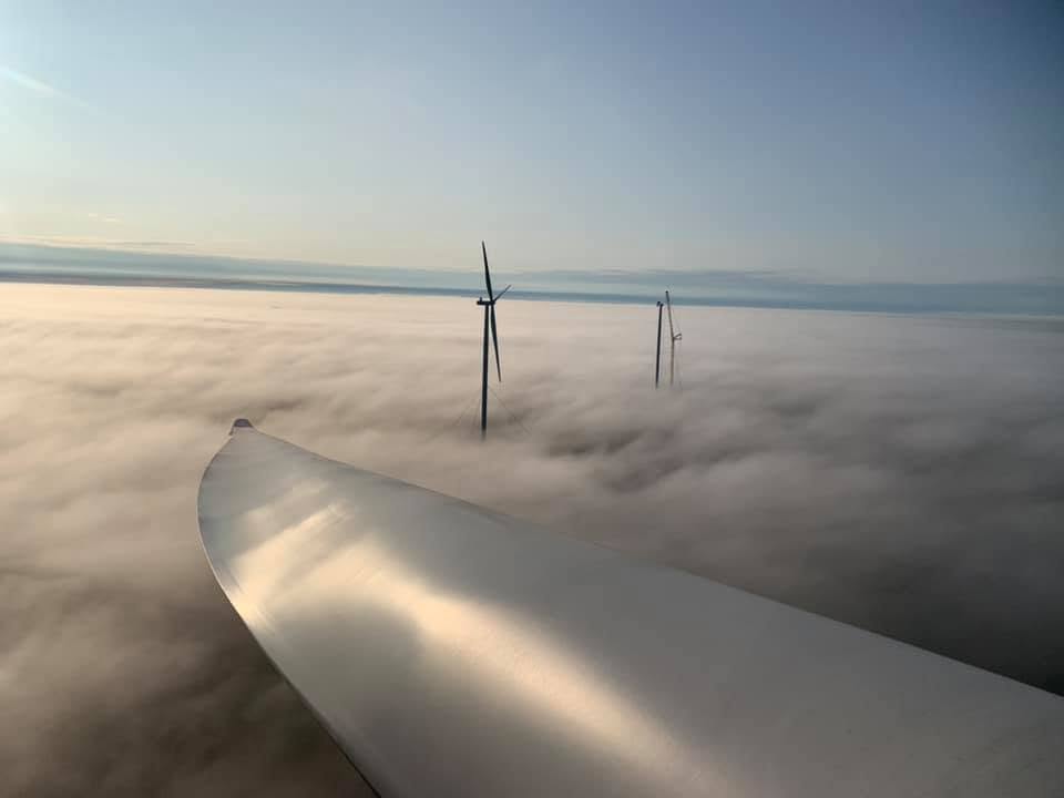 A turbine blade stretching out over low clouds with wind turbines above the mist
